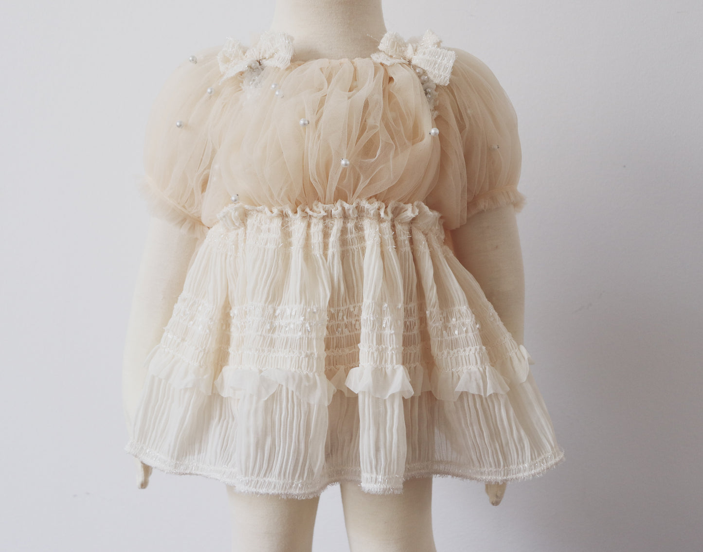 Baby girl wearing an ivory romper with puffy pearl-embellished sleeves and chiffon skirt, sitting sweetly at a baptism photo session