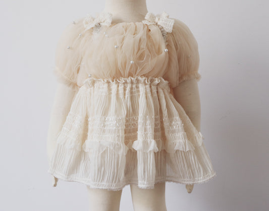 Baby girl wearing an ivory romper with puffy pearl-embellished sleeves and chiffon skirt, sitting sweetly at a baptism photo session