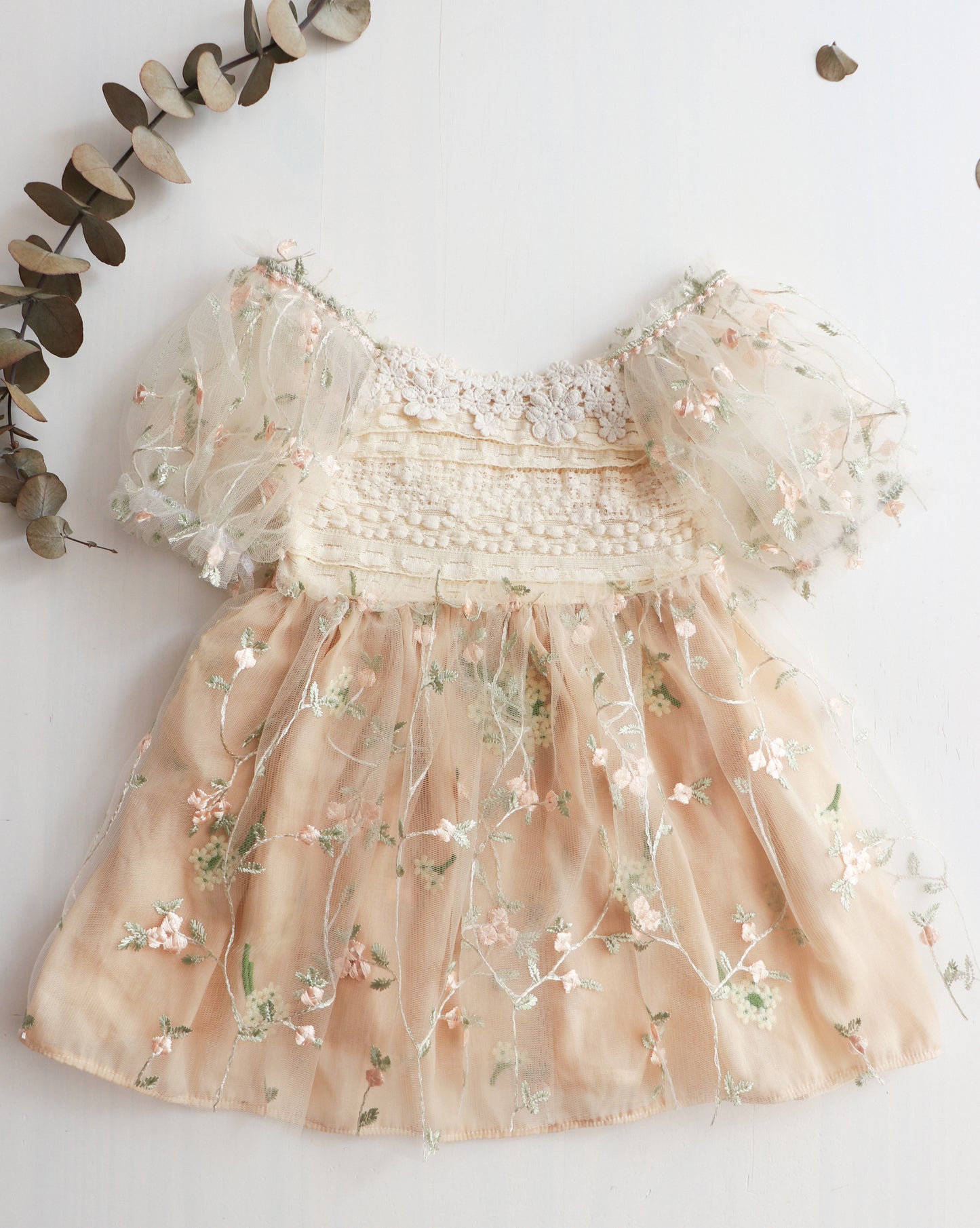 A beige lace dress for girls with embroidered tulle and a floral chiffon skirt, displayed on a white background with decorative leaves.