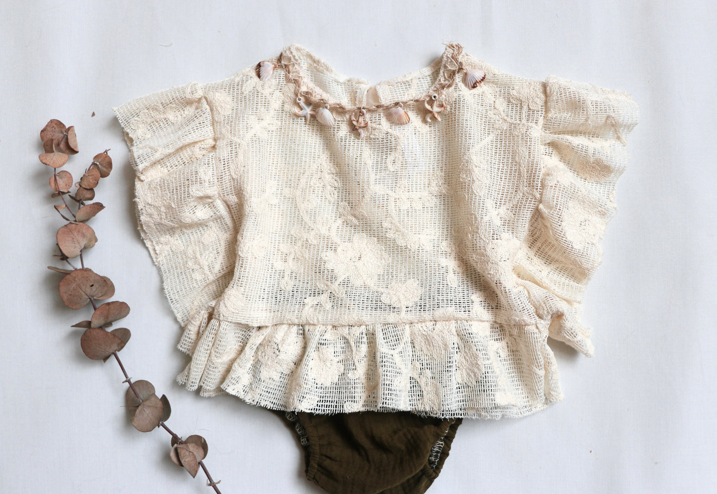 "Baby girl wearing a beige ruffle top with vintage lace and natural shell details, paired with a deep green bloomer during a boho-inspired outdoor session."