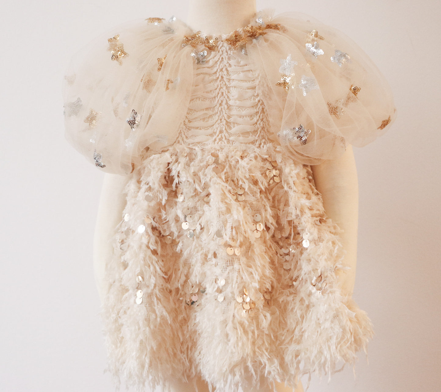 Bring timeless elegance to your child’s photoshoots with this Rococo-inspired girl’s dress. Featuring puffy tulle sleeves adorned with shimmering star sequins, an intricately embroidered fish bone chest, and a flowing light beige feather skirt with gold accents, this dress is perfect for special occasions