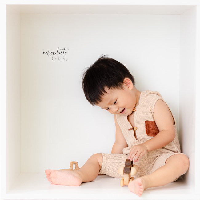Toddler boy wearing beige sitter romper for photoshoot playing with wooden toy inside white box