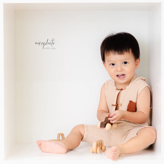 Toddler wearing beige knitted sitter romper for photoshoot holding wooden toy inside white box