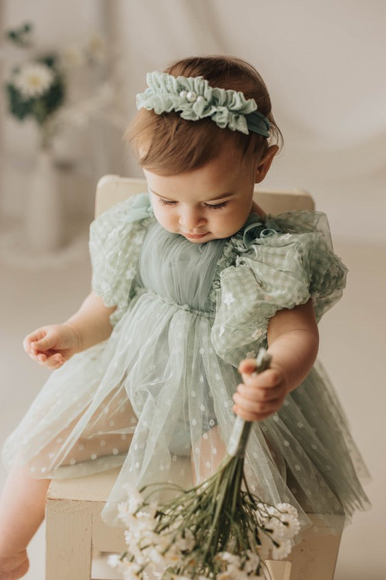Baby girl wearing a sage green baby photo prop dress with puff sleeves and floral headband holding white flowers