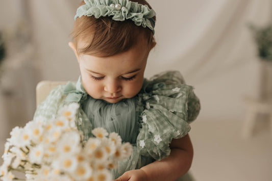 Baby wearing a sage green baby photo prop dress and headband holding a bouquet of daisies
