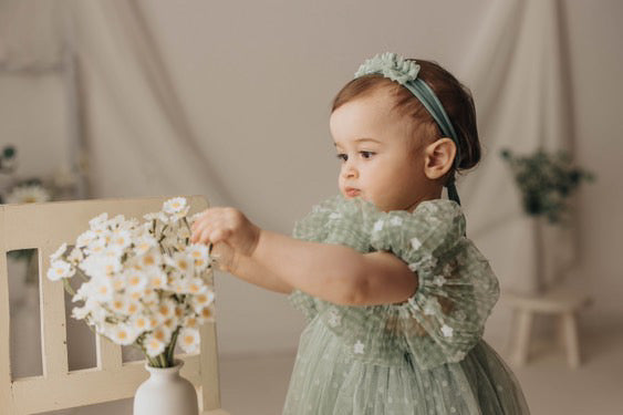Baby girl in sage green dress with matching headband playing with flowers using sage green baby photo prop