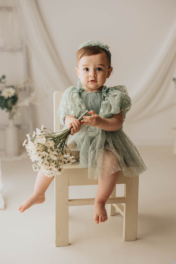 Toddler wearing sage green dress and headband sitting on chair holding flowers with sage green baby photo prop