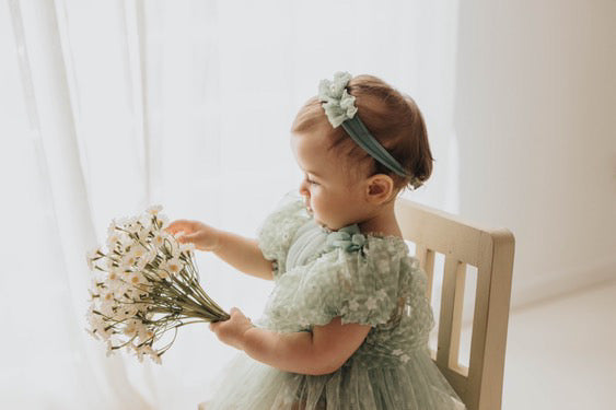 Toddler girl in sage green baby photo prop dress and headband holding white flowers sitting on wooden chair