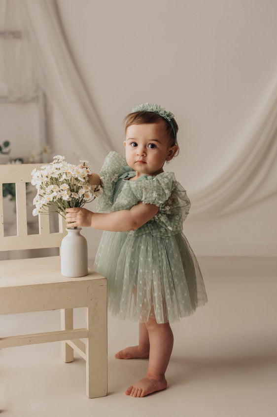 Baby girl in a sage green baby photo prop dress holding a vase of white flowers standing by a small chair