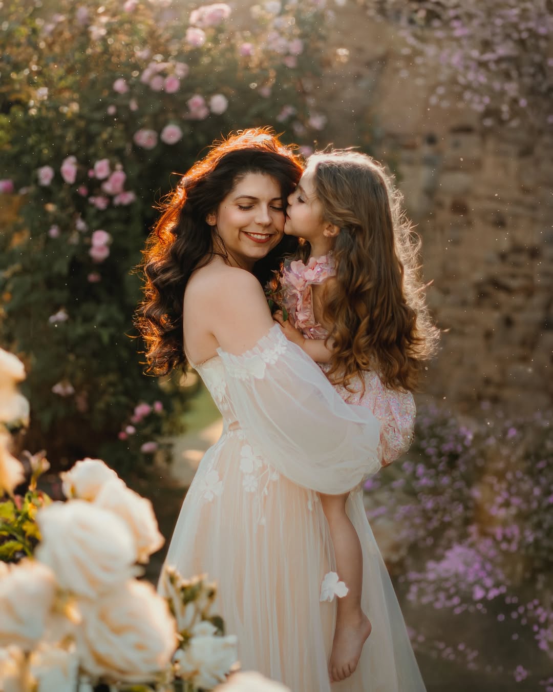 Mother holding daughter wearing sitter session dress pink in a garden with flowers in soft sunlight