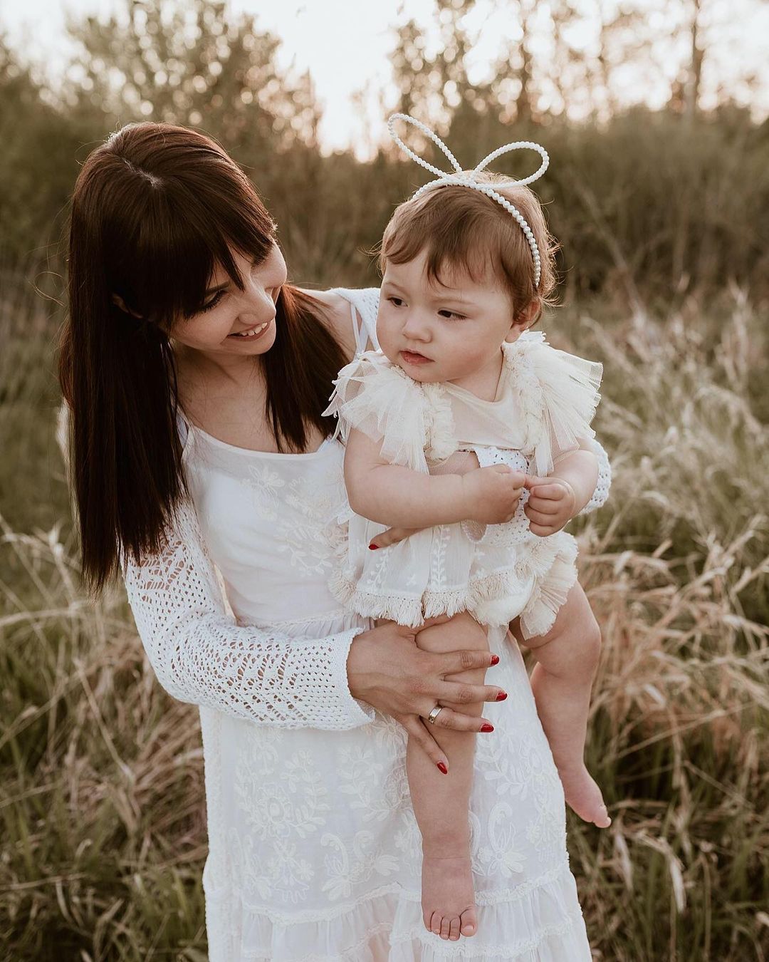 Mother holding baby dressed in delicate white sitter session photography clothes outdoors in natural setting