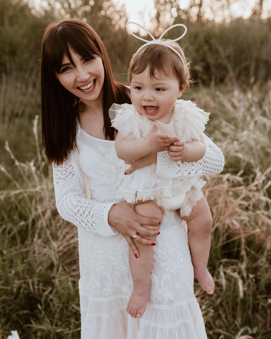Mother holding toddler wearing white fluffy romper and headband in sitter session photography clothes outdoors