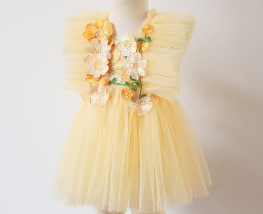 A dreamy girls' playsuit dress in soft yellow cotton with delicate floral embroidery, ruffled tulle on both sides, and crossed adjustable straps, capturing a whimsical flower fairy vibe