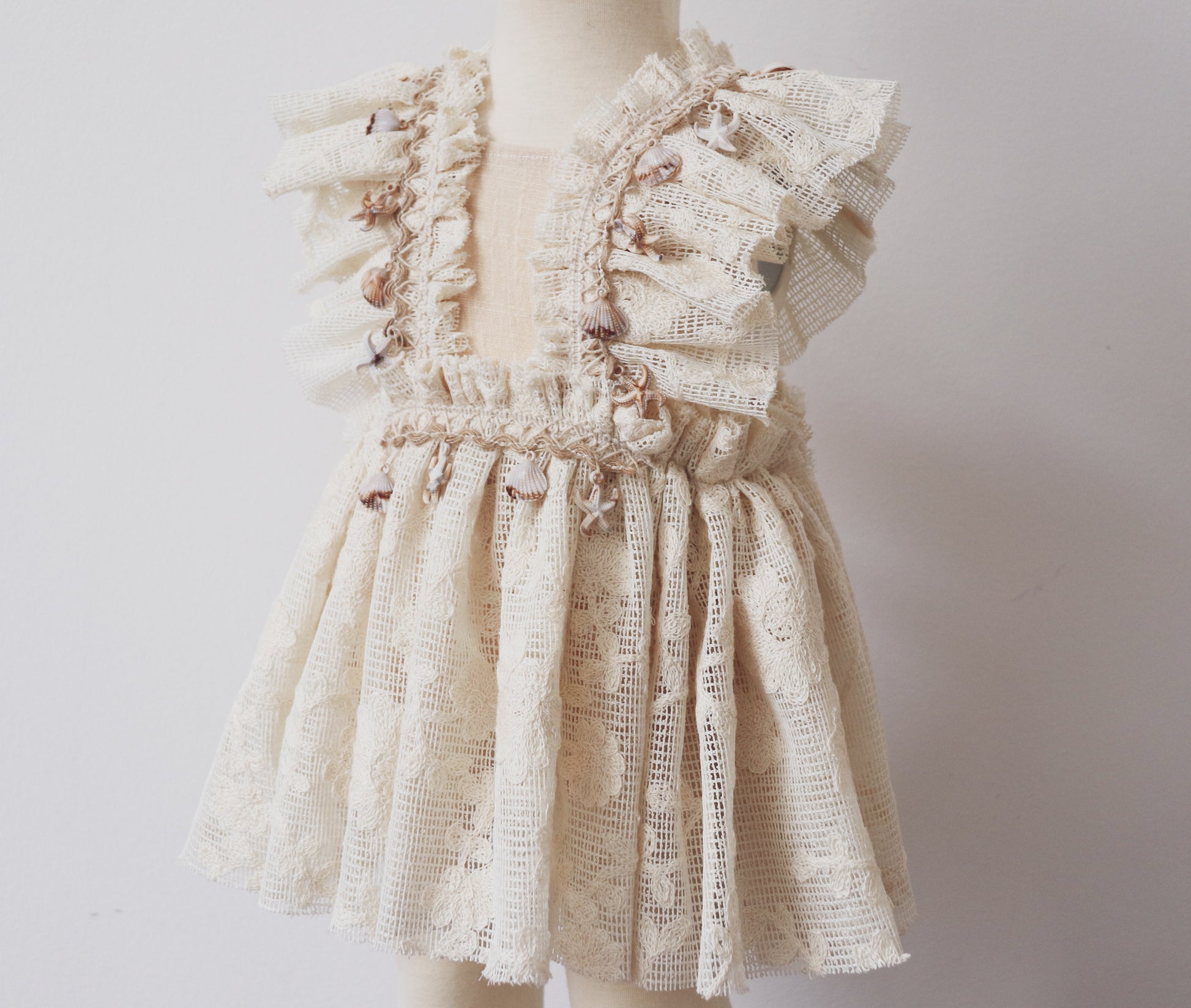 A soft beige cotton playsuit with a vintage overall silhouette, delicate lace accents, and bohemian-inspired fringe with coastal elements, perfect for a relaxed summer look.

