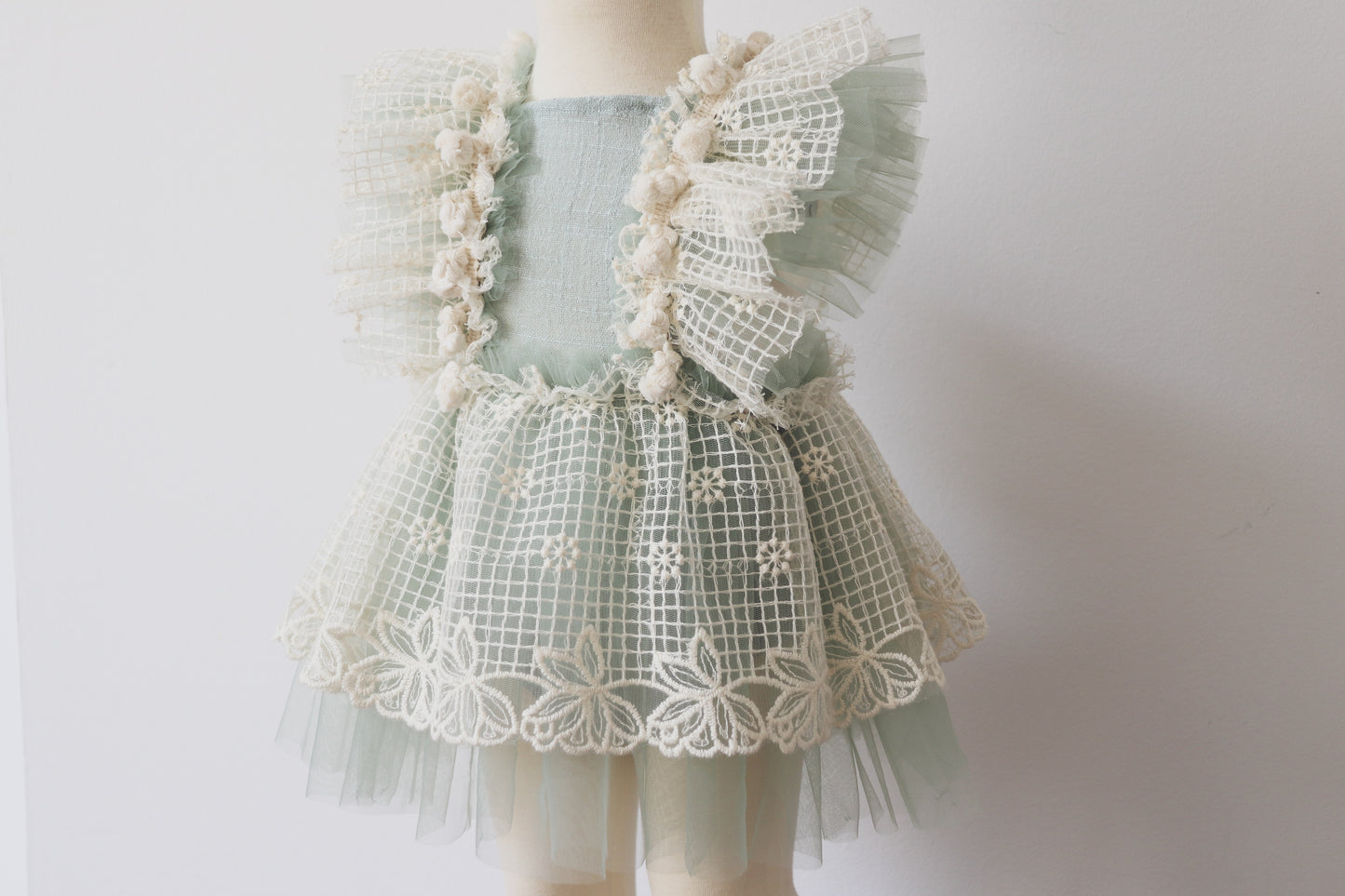 A boho-style girls' playsuit dress in soft sage green cotton, featuring a classic overall silhouette with crossed adjustable straps, vintage beige lace, ruffled sage green tulle, and delicate beige fringes