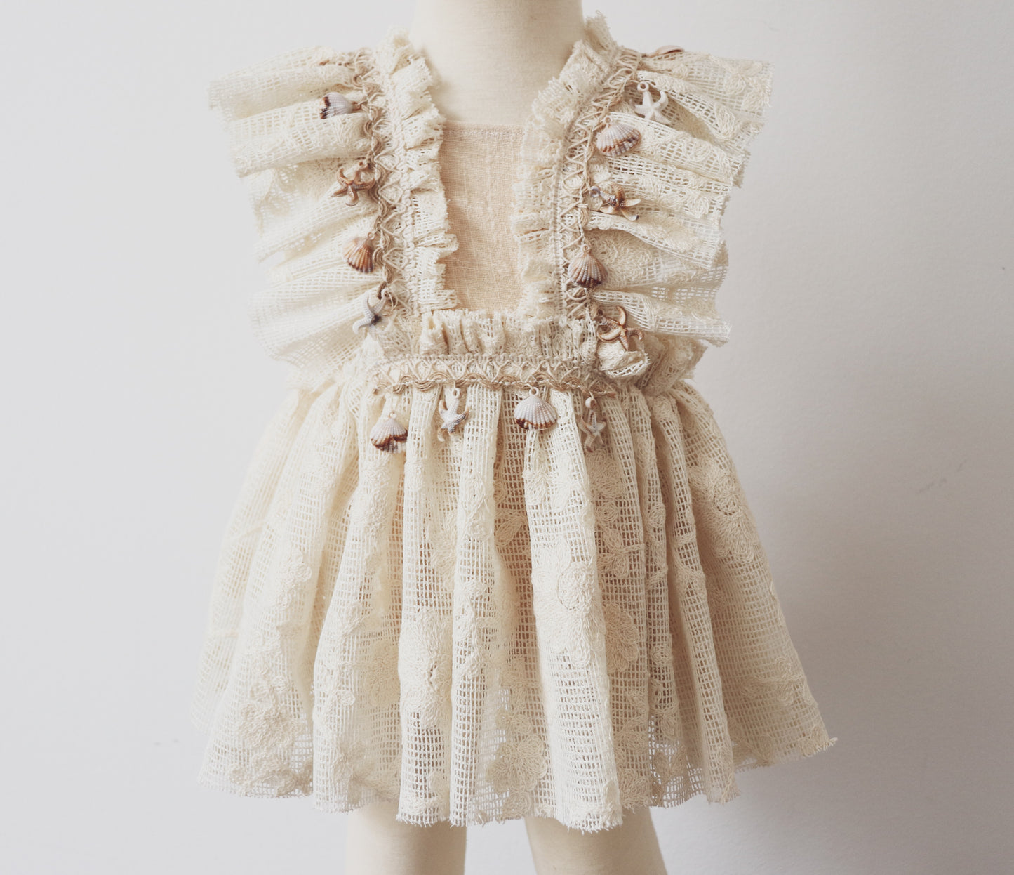 A vintage-inspired girls' playsuit dress in soft beige cotton, featuring ruffled bodice details, vintage lace, and boho-style fringe adorned with seashells and starfish. Perfect for beach photoshoots and bohemian sessions.