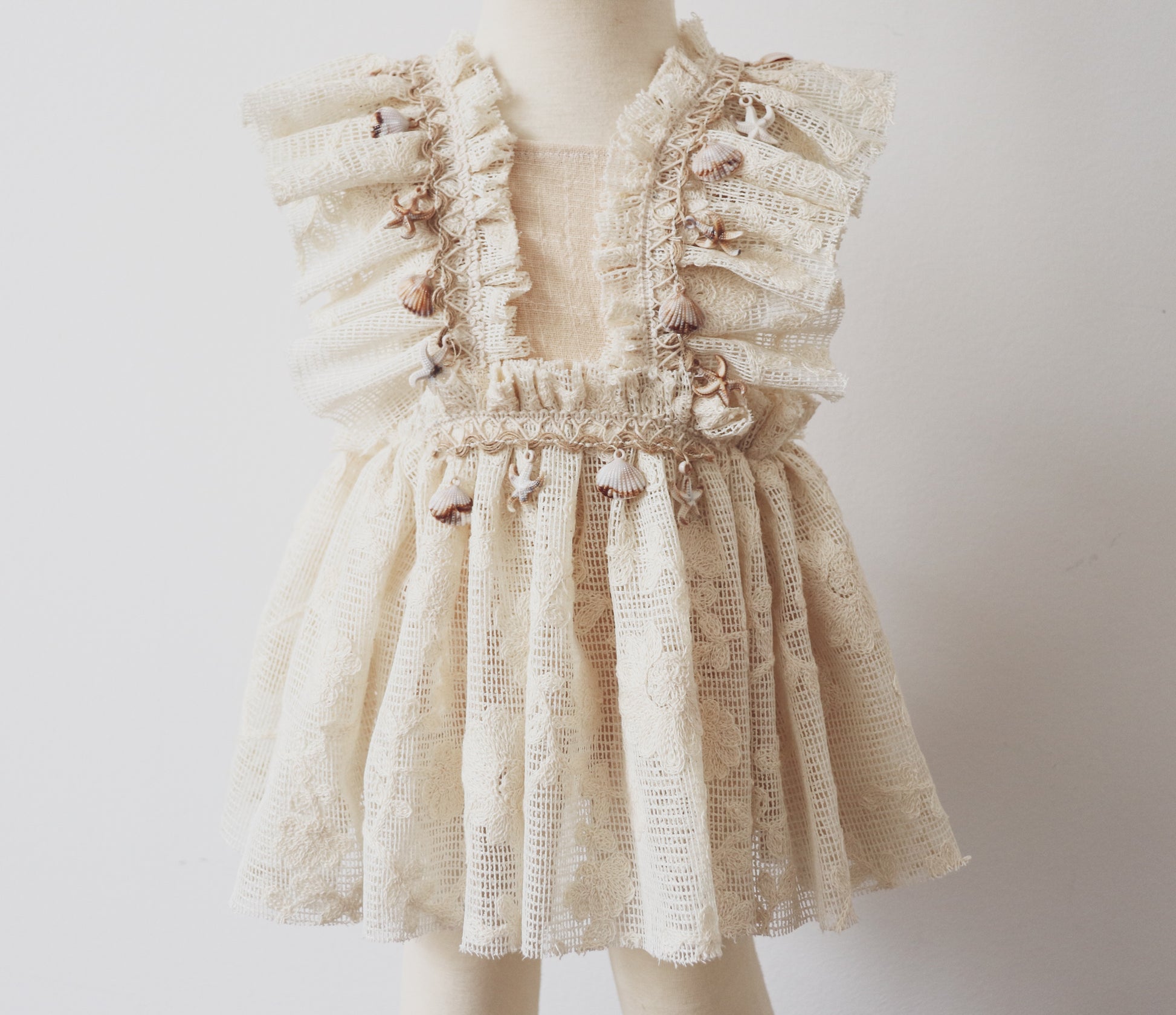 A vintage-inspired girls' playsuit dress in soft beige cotton, featuring ruffled bodice details, vintage lace, and boho-style fringe adorned with seashells and starfish. Perfect for beach photoshoots and bohemian sessions.