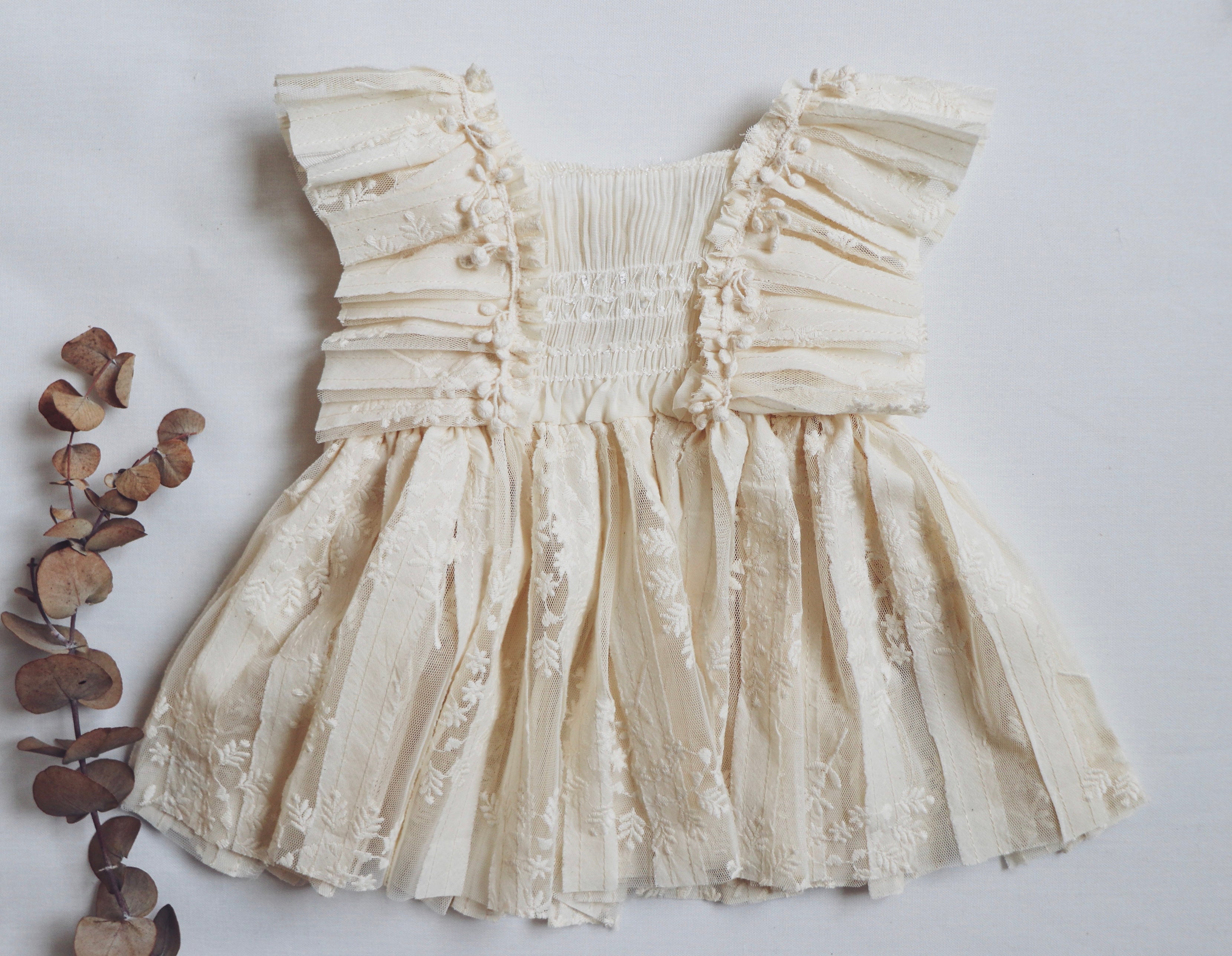Light beige boho-style girl dress with a soft chiffon bodice, intricate floral embroidered lace, sculptural striped lace detailing, and mini cotton pom-poms on both sides. Perfect for photoshoots and special occasions."