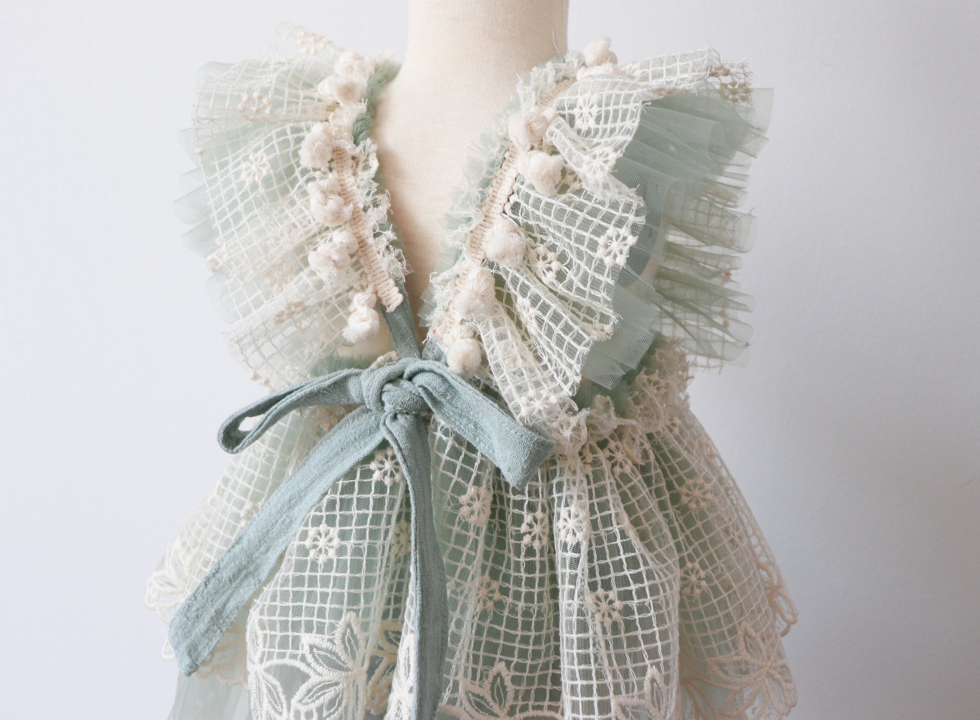 Close-up of a charming girls' overall dress with ruffled sage green tulle, vintage beige lace, and soft beige fringes, creating a whimsical boho look