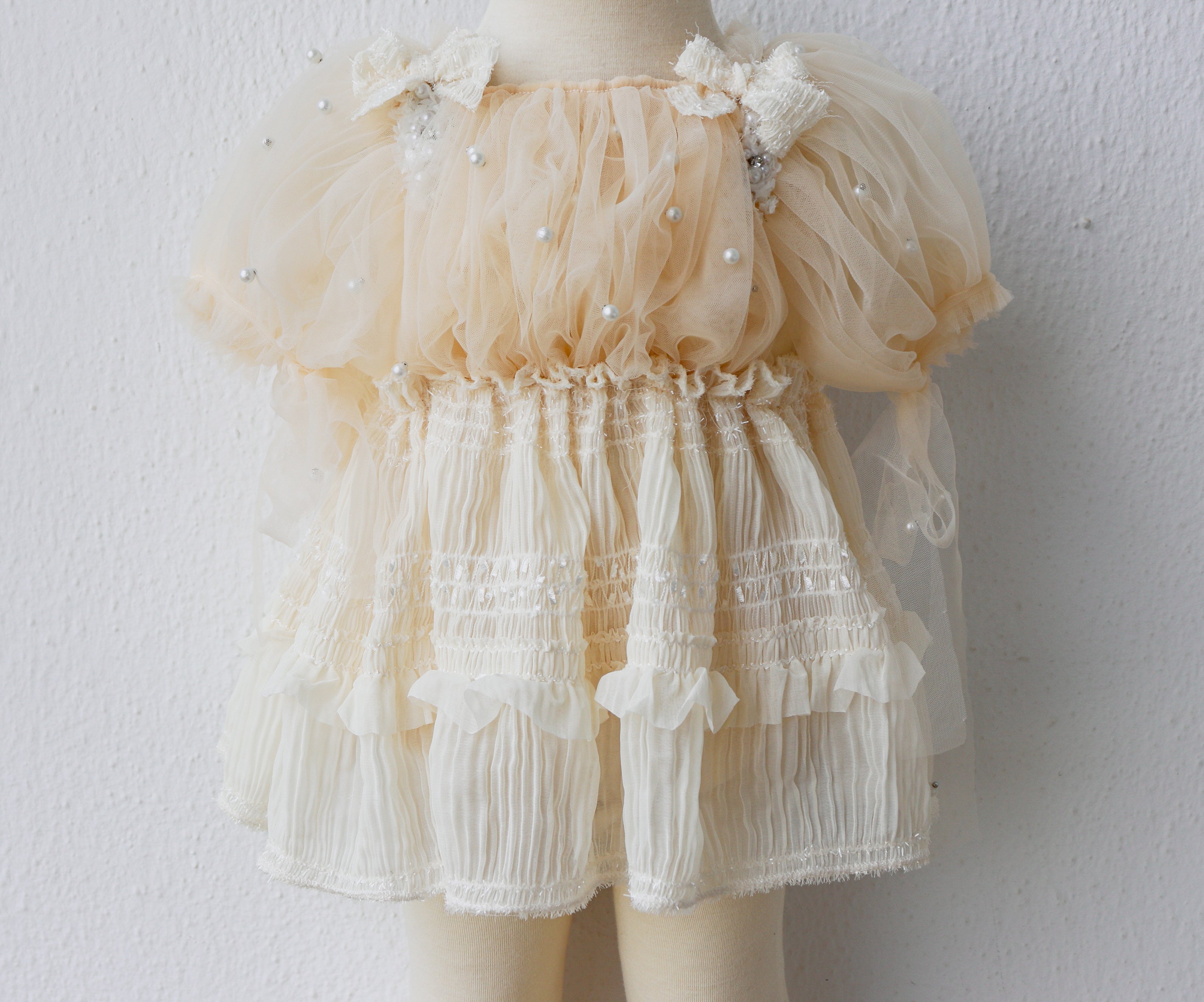 Baby girl wearing an ivory romper with puffy pearl-embellished sleeves and chiffon skirt, sitting sweetly at a baptism photo session