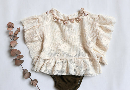 "Baby girl wearing a beige ruffle top with vintage lace and natural shell details, paired with a deep green bloomer during a boho-inspired outdoor session."