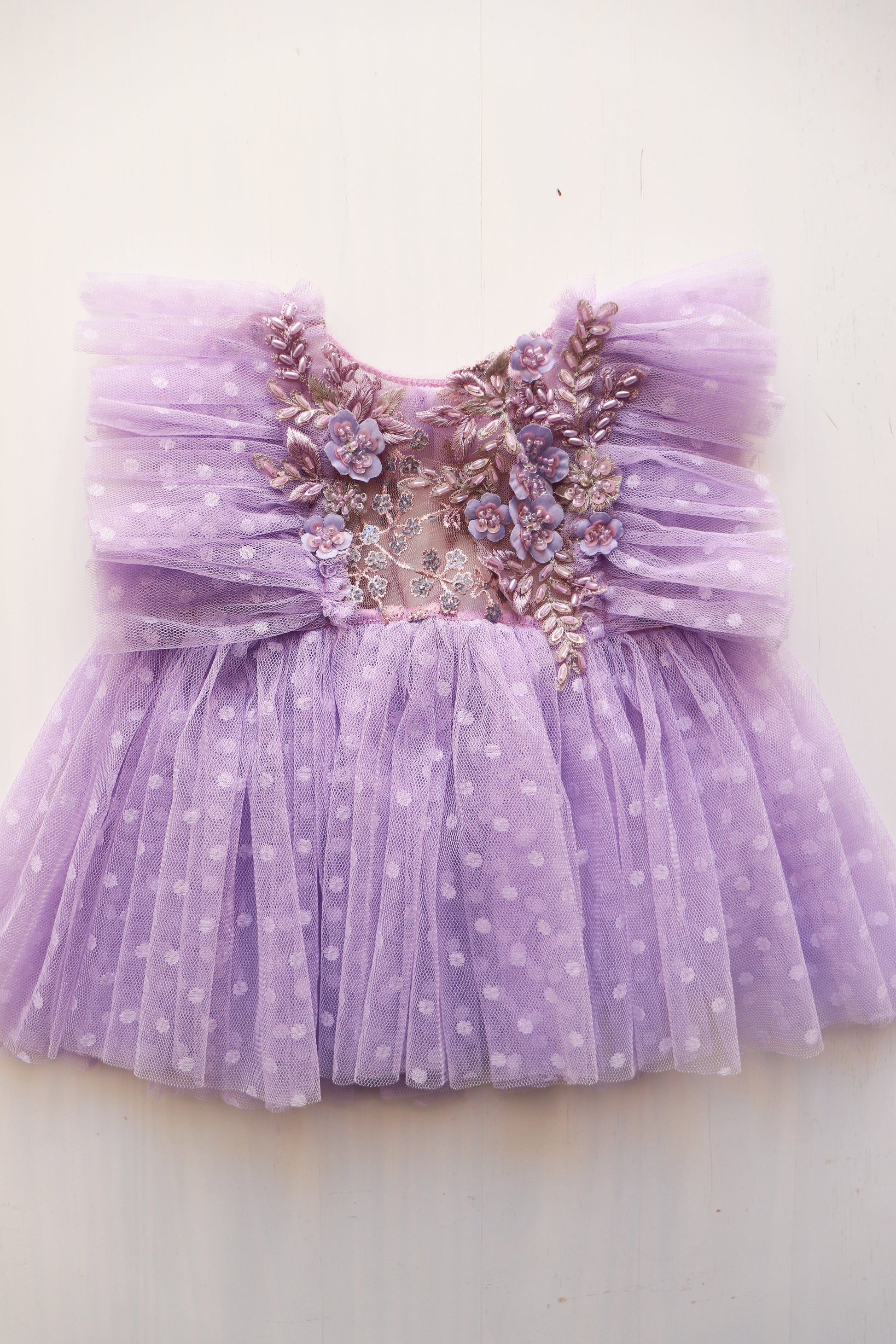 Inspired by nature’s beauty, this dress features delicate 3D flowers, pearls, and a Hortensia tieback in soft lavender. The dotted tulle overlay and matching diaper cover complete the dreamy, boho-inspired look.

