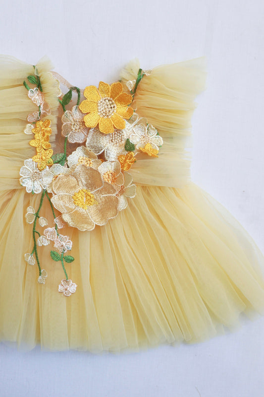 "Little girl in a pastel yellow tulle dress with 3D embroidered flowers and ruffled sides, smiling during a sunny outdoor photo session."

