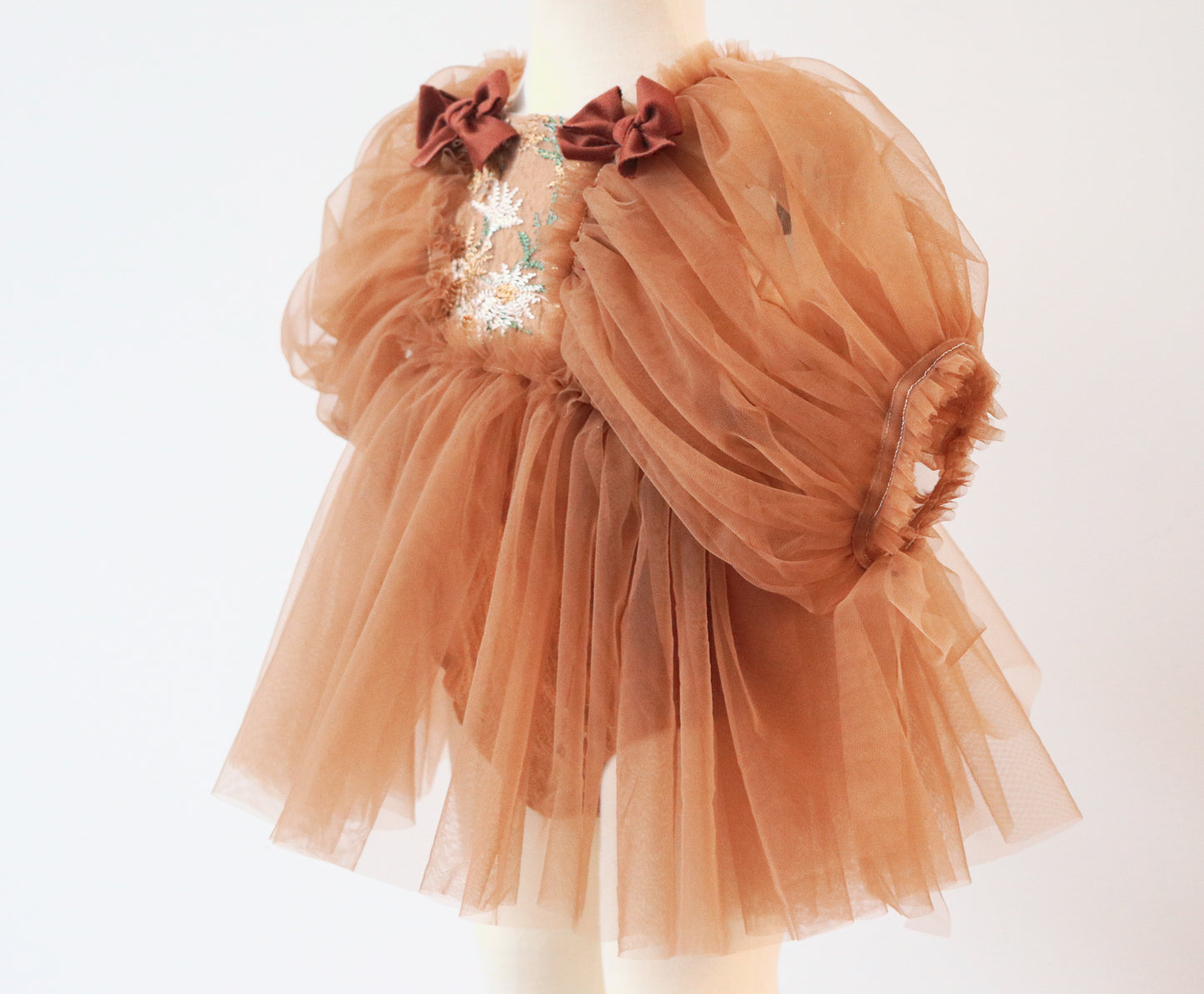 "Close-up of a girl’s dress featuring a bronze embroidered bodice, soft brown tulle skirt, and puffy tulle sleeves with velvet bow details."

