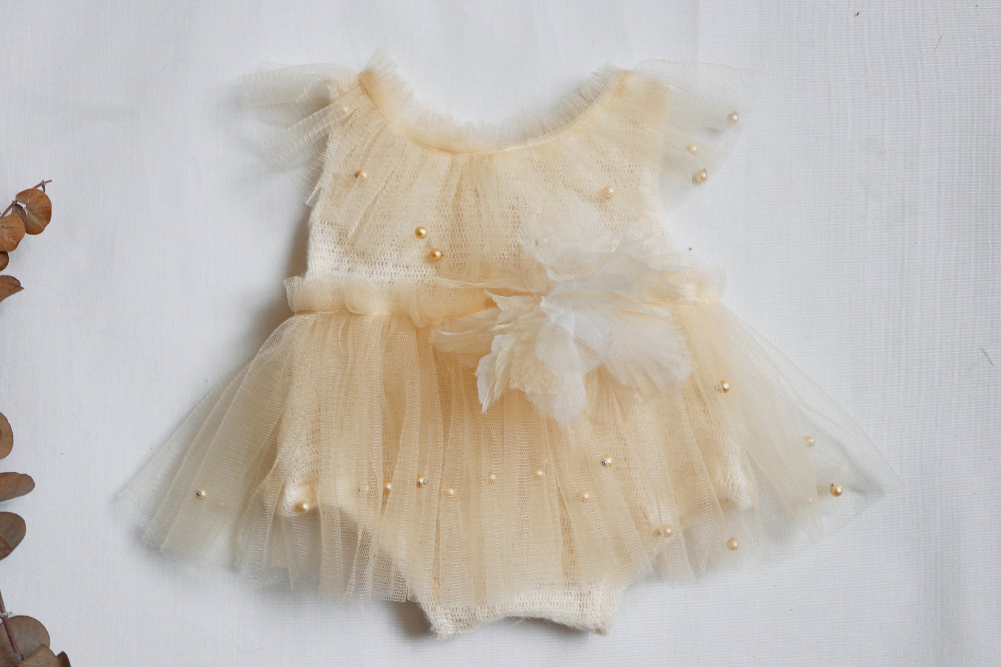 "Newborn girl wearing a soft cream romper dress with champagne tulle, pearl accents, and a chiffon flower detail during a delicate studio photo session."

