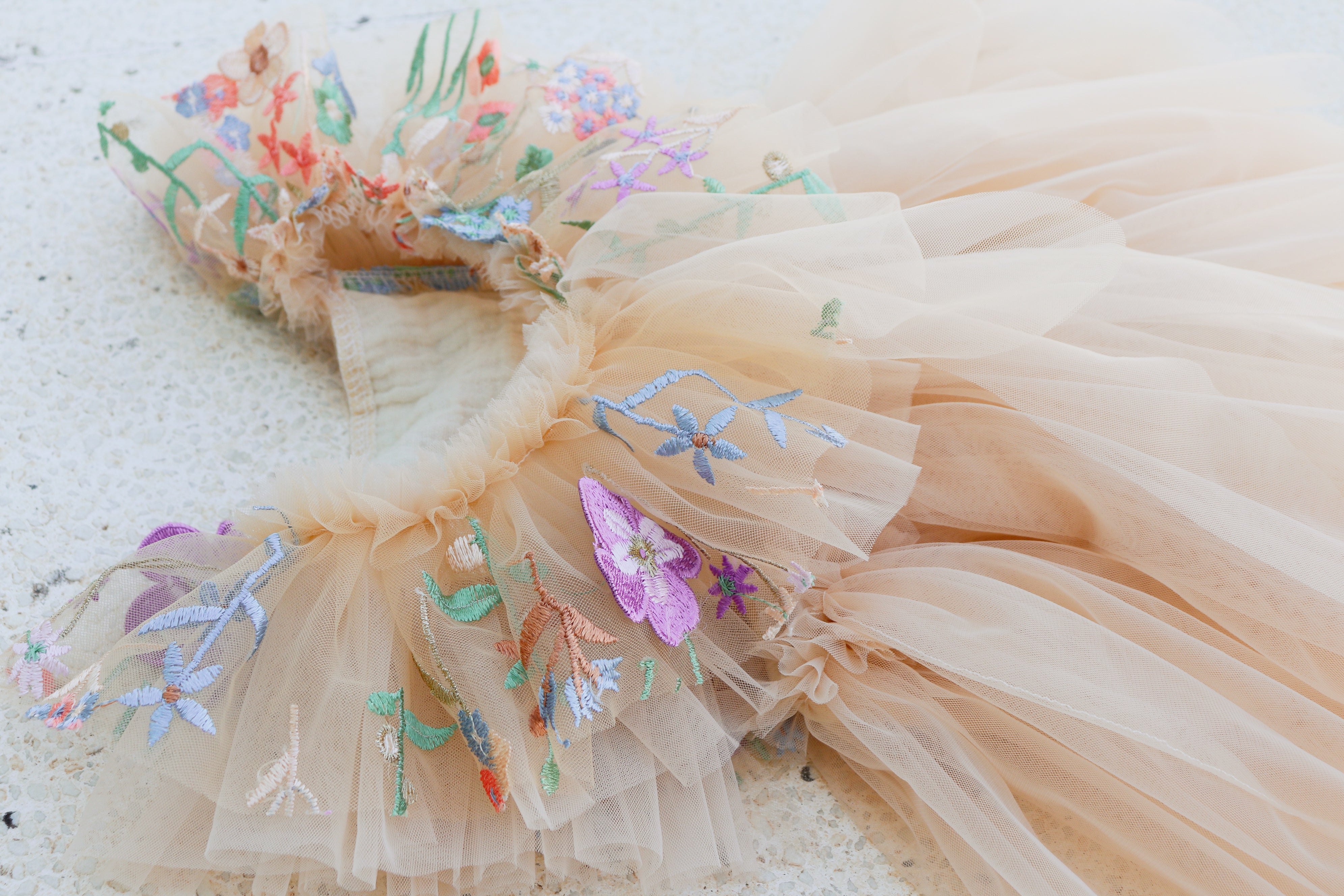 Close-up of nude tulle girl dress with delicate floral embroidery and soft ruffles, styled for a natural outdoor shoot in golden light.

