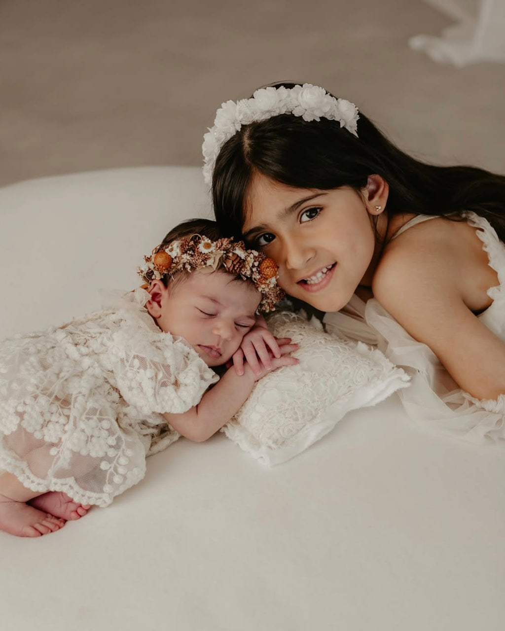 Newborn baby wearing lace newborn photo outfit sleeping beside smiling older sibling with flower headbands