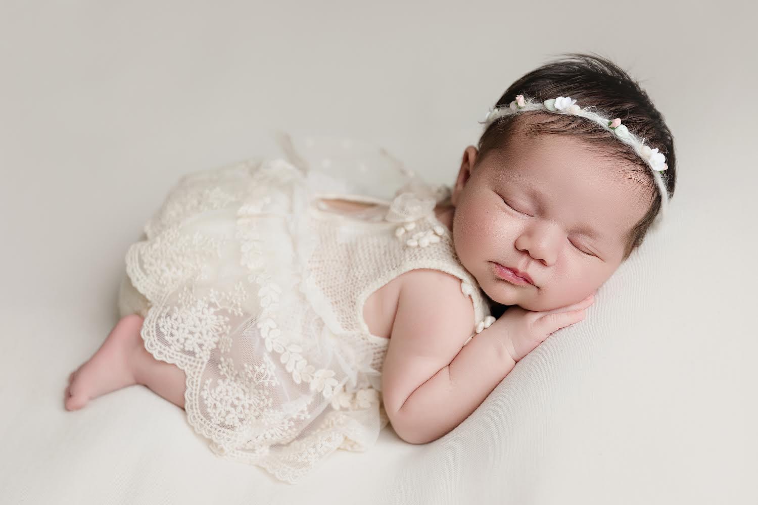 Sleeping baby wearing a white newborn lace romper outfit with floral headband on a soft surface